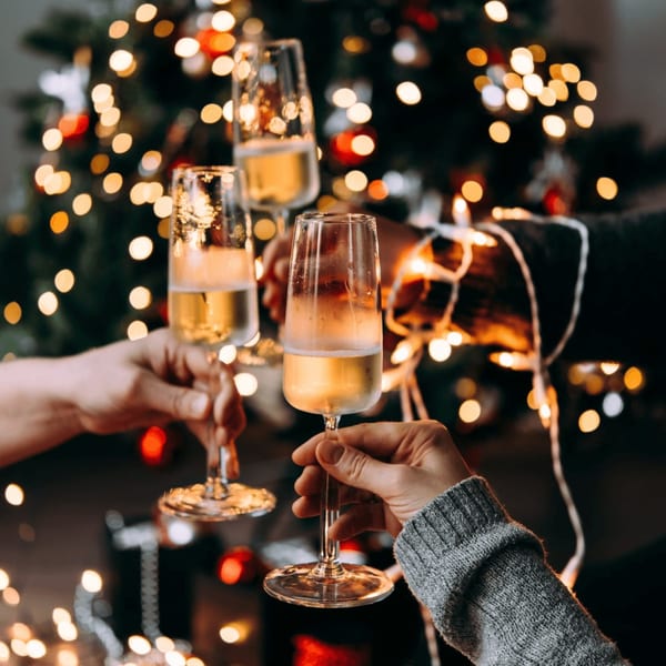 Three hands clinking champagne flutes in front of a festive, blurred Christmas tree with lights.