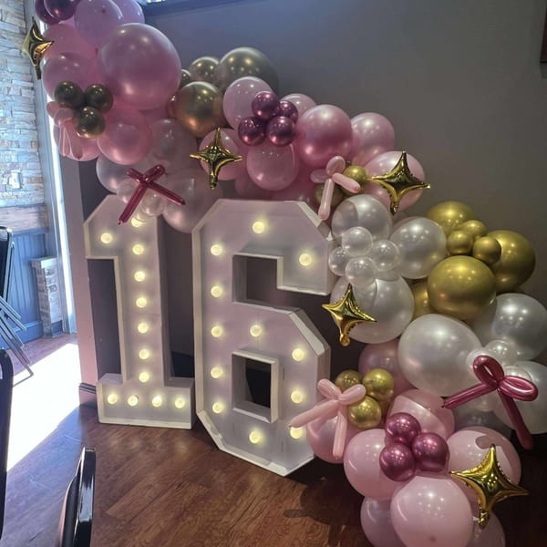 Lighted "16" sign surrounded by pink, gold, and white balloons, with star-shaped accents.