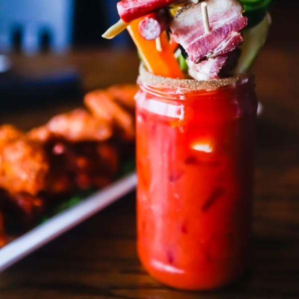 A Bloody Mary cocktail in a mason jar, garnished with bacon, celery, and peppers, next to chicken wings.