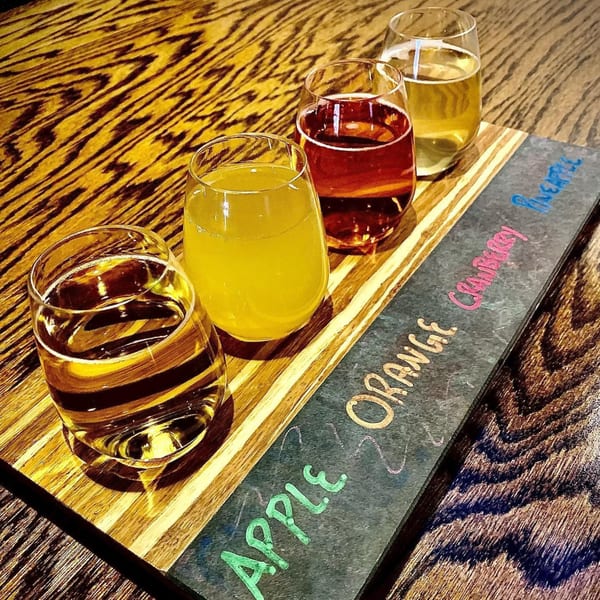 Four cider samples on a wooden paddle, labeled Apple, Orange, Cranberry, and Pineapple.